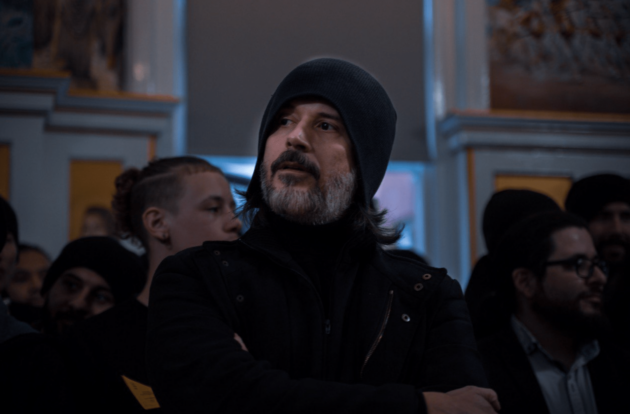 Detailný záber na Aba Al-Sadiqa s prekríženými rukami na hrudi a vážnym, odhodlaným pohľadom smerujúcim nahor, mimo svetské dianie. Toto gesto symbolizuje duchovný odpor, kategorické odmietnutie účasti na voľbách 'menšieho zla' a vernosť výhradne Božiemu vedeniu.